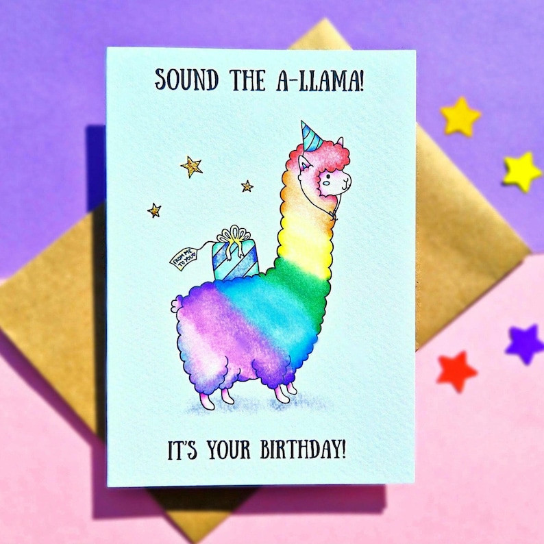 Puede incluir: Una tarjeta de cumplea&ntilde;os con un dibujo animado de llama que lleva un gorro de fiesta y lleva un regalo. La llama es de color arco&iacute;ris y la tarjeta dice "Sound the A-llama! It's your birthday!"