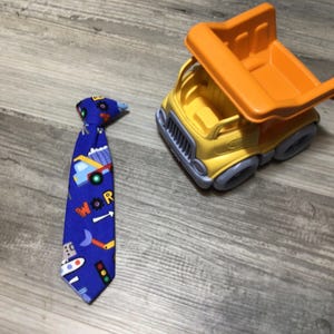 May include: A blue tie with a colorful transportation-themed pattern, including trucks, cars, and the word "WOR". Beside it is a yellow and orange toy dump truck, perfect for a child's playtime.