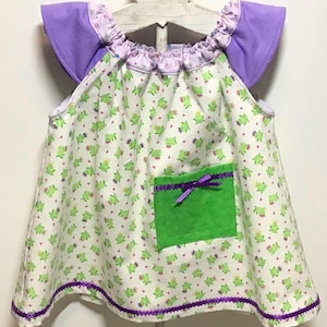 May include: A white baby dress with a green frog print and purple accents. The dress features a green pocket with a purple bow, purple trim, and purple flutter sleeves. The dress is hanging on a wooden hanger.