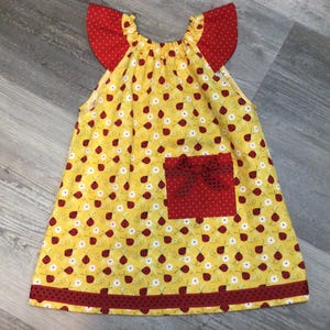 May include: A yellow sleeveless dress with a ladybug pattern, red polka dot accents, and a red bow pocket. The dress has red polka dot trim and red ruffled sleeves.
