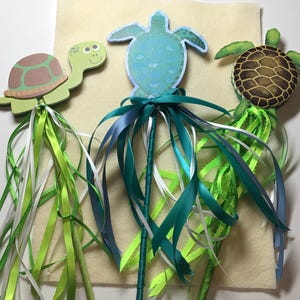 May include: Three wooden turtle wands with colorful ribbons. The turtles have green, brown, and blue shells. The ribbons are shades of green, blue, and white. These wands are suitable for children's parties or as decorative items.