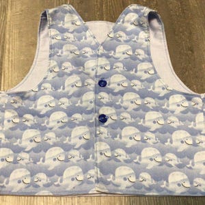 May include: A light blue vest with a repeating pattern of smiling white whales on a blue background. The vest has a V-neck, two blue buttons, and a light purple lining.