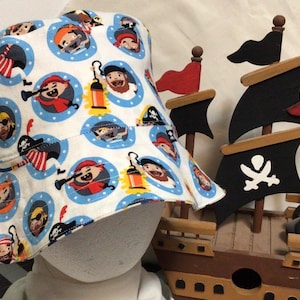 May include: White bucket hat with a repeating pattern of cartoon pirates in blue circles. A wooden pirate ship model with black sails and red flags is in the background. The hat is a fun accessory for a child.