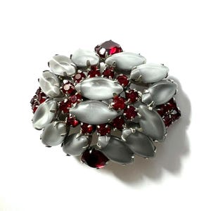 May include: A cluster brooch featuring a dense arrangement of oval-shaped, milky white cabochon stones interspersed with small, faceted deep red crystals. The stones are set in a silver-toned metal frame, creating a layered, textured floral appearance.