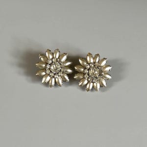 May include: A pair of vintage-style clip-on earrings. Each earring is shaped like a flower with a central clear rhinestone surrounded by faux pearls. The petals are pearlescent, and the center stone sparkles. The earrings are set against a gray background.