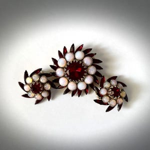May include: Three pieces of jewelry with a floral design. Each piece features a central red stone surrounded by white and red gems. The pieces are arranged in a line on a white surface.