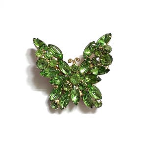 May include: A green butterfly brooch with a gold-tone metal frame. The brooch is covered in various shades of green rhinestones, creating a sparkling effect. The butterfly's wings are detailed with marquise-cut stones.
