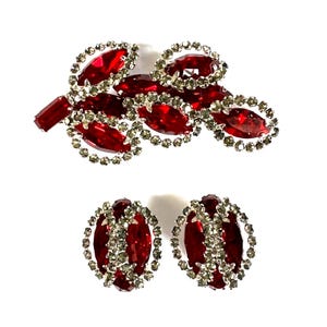 May include: A set of vintage jewelry, including a brooch and a pair of clip-on earrings. The brooch features a cluster of red oval-shaped stones with a silver-colored border. The earrings have a similar design, with red stones and a silver-colored border.