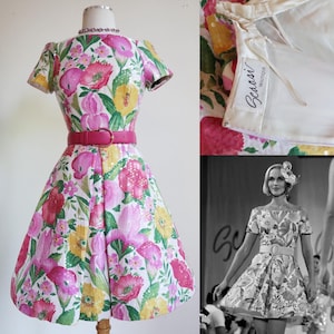 Puede incluir: Un vestido blanco con estampado floral rosa, amarillo y verde. El vestido tiene un cinturón rosa y mangas cortas. El vestido está etiquetado como "Bessoni Boutique".