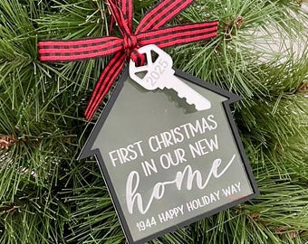 May include: A house-shaped Christmas ornament in olive green with a black border, featuring a white key cutout labeled 2025. The text reads FIRST CHRISTMAS IN OUR NEW home and 1944 HAPPY HOLIDAY WAY. It is tied with a red and black buffalo plaid ribbon.