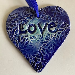 May include: A blue ceramic heart-shaped ornament with a white glaze and a floral pattern. The word "Love" is embossed on the front of the ornament.