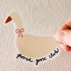May include: A die-cut sticker featuring a cartoon goose with a light beige body, a red-brown beak, and a pink bow. The text "porch goose club" is written in black script along the bottom edge of the sticker.