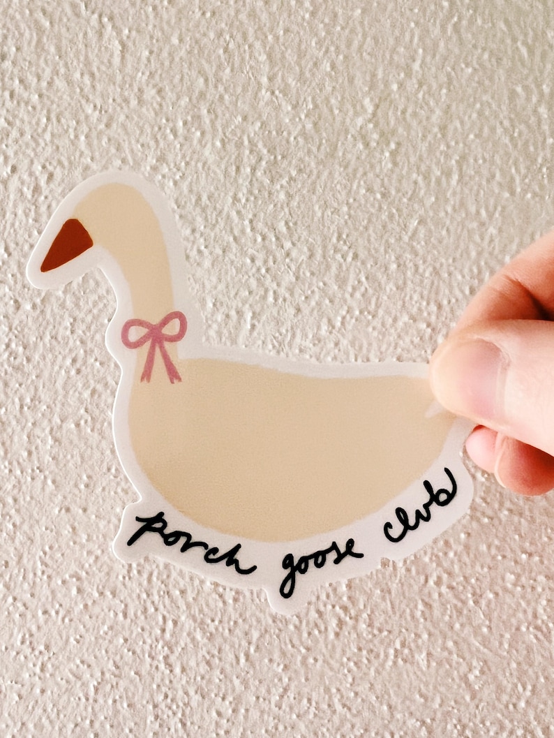 May include: A die-cut sticker featuring a cartoon goose with a light beige body, a red-brown beak, and a pink bow. The text "porch goose club" is written in black script along the bottom edge of the sticker.