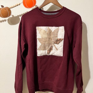 Op de afbeelding: Een bordeauxrood sweatshirt met een patchwork-bladontwerp op de voorkant. Het blad is gemaakt van verschillende stoffen in tinten bruin, beige en wit.