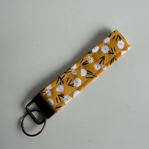 May include: Mustard yellow fabric key fob with a white floral pattern and black accents. The key fob has a dark brown loop and a metal key ring. The key fob is approximately 6 inches long.