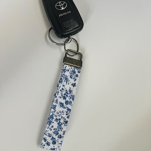 May include: A black Toyota RAV4 key fob attached to a key ring and a fabric wristlet. The wristlet is white with a blue floral pattern and a bronze-colored metal clasp.