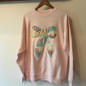 May include: A light pink long-sleeve sweatshirt featuring a large, centered fabric bow appliqué. The bow is constructed from various patchwork fabric segments in shades of teal, yellow, and white, decorated with floral and striped patterns.