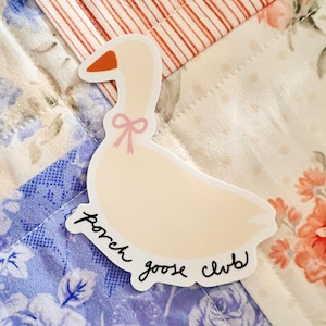 May include: A die-cut sticker featuring a cartoon goose with a pink bow and orange beak. The sticker has the words "porch goose club" in a handwritten font. The sticker is placed on a floral patterned quilt.