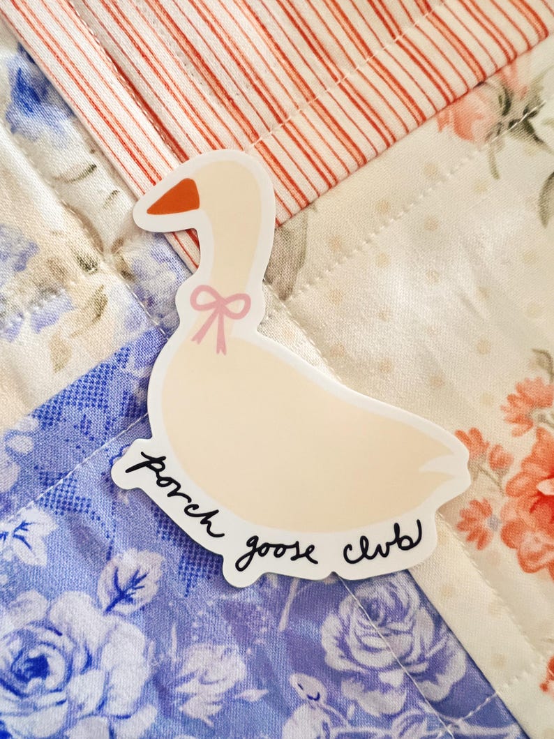 May include: A die-cut sticker featuring a cartoon goose with a pink bow and orange beak. The sticker has the words "porch goose club" in a handwritten font. The sticker is placed on a floral patterned quilt.