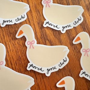 May include: Cream-colored goose stickers with orange beaks and pink bows around their necks. The stickers have the words "porch goose club" in a handwritten font. The stickers are on a wooden surface.