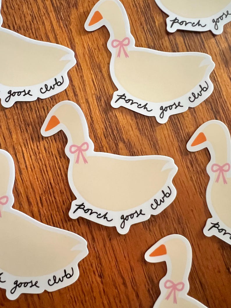 May include: Cream-colored goose stickers with orange beaks and pink bows around their necks. The stickers have the words "porch goose club" in a handwritten font. The stickers are on a wooden surface.