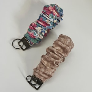 May include: Two fabric wristlets with metal hardware. One wristlet features a floral pattern in pink, blue, and green. The other wristlet is a solid tan color with a subtle floral design. Both have a gathered, ruched appearance.