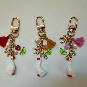以下が含まれることがあります： ゴールドカラーの金具が付いた、3つの装飾的なキーホルダー。各キーホルダーには、赤いアクセントが付いた白いガチョウのチャームがあり、タッセル、ビーズ、さまざまな色の花のチャームで飾られています。魅力的なアクセサリー。