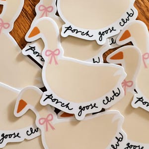 May include: A collection of stickers featuring a cartoon goose design. The geese are cream-colored with orange beaks and a pink bow. The text "porch goose club" is written in a cursive font around the goose's body.