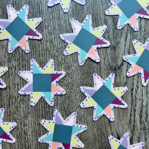 May include: A collection of colorful star-shaped stickers. Each sticker features a geometric design with teal, purple, yellow, and pink segments. The edges are decorated with small blue floral patterns, creating a patchwork quilt aesthetic.