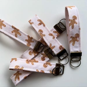 May include: Gingerbread man wristlet keychains. These keychains feature a white background with a repeating pattern of gingerbread men. Each keychain has a bronze-colored metal clasp and ring.