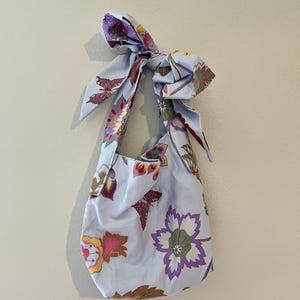 May include: A light blue fabric shoulder bag with a floral and butterfly pattern. The bag has a bow at the top and a matching strap. The flowers are purple, yellow, and brown. The bag is hanging against a light beige wall.