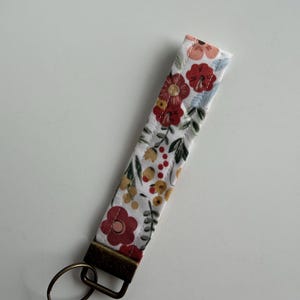 May include: A floral wristlet key fob with a white background and a pattern of red, yellow, and pink flowers. The key fob has a bronze-colored metal clasp and key ring.