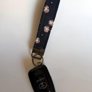 May include: A black key wristlet with a floral pattern in light brown and white, attached to a black Toyota RAV4 key fob. The wristlet has a bronze-colored metal clasp and ring.