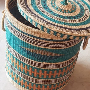 Op de afbeelding: Een geweven opbergmand met een deksel. De mand is gemaakt van natuurlijke materialen en heeft een gestreept patroon in turquoise, bruin en oranje. Het deksel heeft een cirkelvormig patroon in turquoise en bruin.