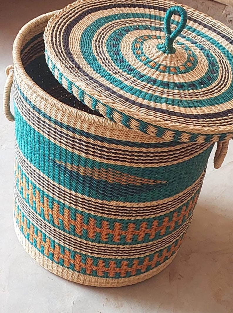 Op de afbeelding: Een geweven opbergmand met een deksel. De mand is gemaakt van natuurlijke materialen en heeft een gestreept patroon in turquoise, bruin en oranje. Het deksel heeft een cirkelvormig patroon in turquoise en bruin.