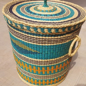 Op de afbeelding: Een geweven opbergmand met een deksel en handvatten. De mand is gemaakt van natuurlijke materialen en heeft een blauw, oranje en bruin patroon.