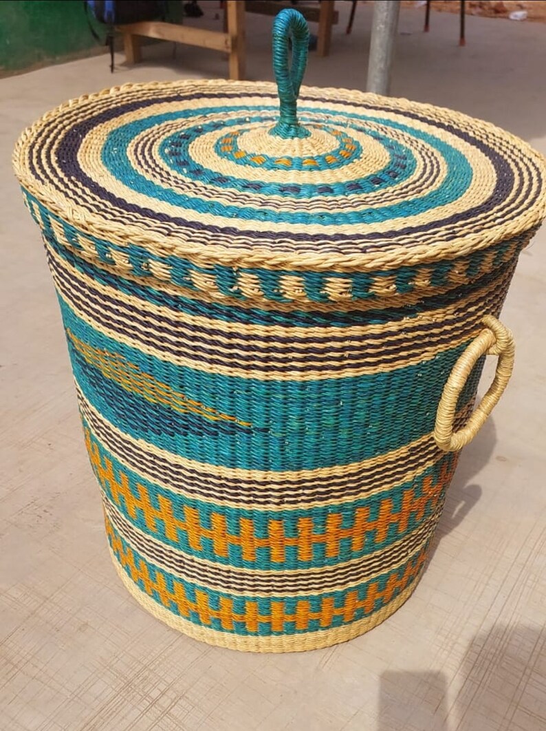 Op de afbeelding: Een geweven opbergmand met een deksel en handvatten. De mand is gemaakt van natuurlijke materialen en heeft een blauw, oranje en bruin patroon.