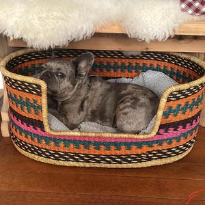 May include: A gray and white French bulldog dog laying in a woven oval pet bed with a colorful geometric pattern. The bed is made of natural materials and has a soft, comfortable lining.