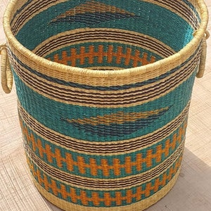 Op de afbeelding: Een geweven opbergmand met een gestreept patroon in turquoise, bruin en oranje. De mand heeft twee handvatten en is gemaakt van natuurlijke materialen.
