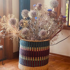 May include: A large, woven basket with a colorful striped pattern in shades of green, brown, and purple. The basket is filled with dried flowers, including thistle and globe thistle.