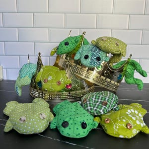 May include: Handmade plush frog toys in shades of green, with different patterns and button eyes. Some frogs are arranged on and around a gold decorative crown.