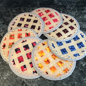 May include: Eight fabric pie-shaped coasters with different fruit designs. The coasters are beige with a woven texture and feature a variety of fruits, including blueberries, cherries, peaches, and strawberries.