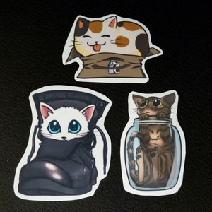 May include: Three stickers featuring cartoon cats in different poses. One cat is in a brown paper bag, another is in a glass jar, and the third is in a black boot.