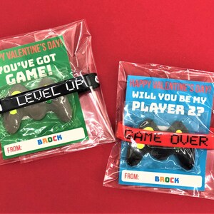 May include: Two Valentine's Day cards with a video game theme. The cards are packaged in clear plastic bags. The first card says "Happy Valentine's Day! You've Got Game!" and features a black wristband with the text "Level Up". The second card says "Happy Valentine's Day! Will You Be My Player 2?" and features a red wristband with the text "Game Over". Both cards feature a plastic video game controller.