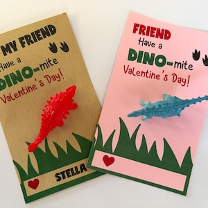May include: Two Valentine's Day cards with dinosaur toys. The card on the left is brown with green grass and the text "To My Friend Have a Dino-mite Valentine's Day!" The card on the right is pink with green grass and the text "Friend Have a Dino-mite Valentine's Day!"