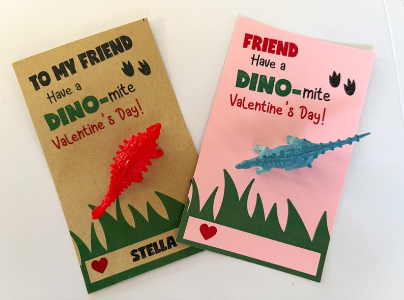 May include: Two Valentine's Day cards with dinosaur toys. The card on the left is brown with green grass and the text "To My Friend Have a Dino-mite Valentine's Day!" The card on the right is pink with green grass and the text "Friend Have a Dino-mite Valentine's Day!"