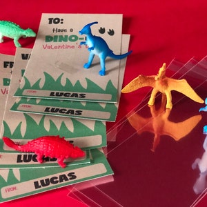 May include: A set of four brown cards with green grass and the text "TO: Have a DINO-Valentine's" and "FROM: LUCAS". The cards are stacked on top of each other. There are five small plastic dinosaur toys in the image, including a green dinosaur, a blue dinosaur, a red dinosaur, a yellow dinosaur, and a blue dinosaur.