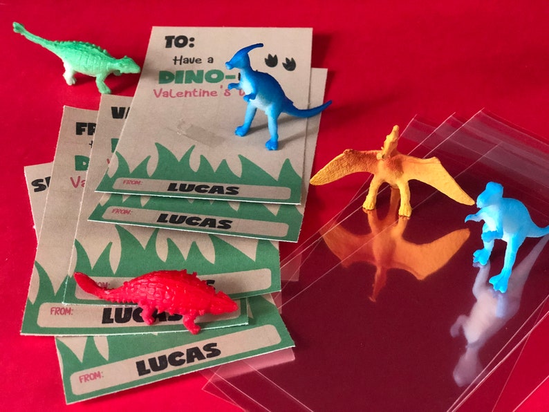 May include: A set of four brown cards with green grass and the text "TO: Have a DINO-Valentine's" and "FROM: LUCAS". The cards are stacked on top of each other. There are five small plastic dinosaur toys in the image, including a green dinosaur, a blue dinosaur, a red dinosaur, a yellow dinosaur, and a blue dinosaur.