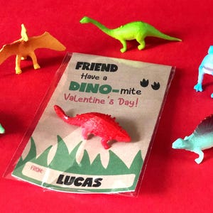 May include: A Valentine's Day card with a dinosaur theme. The card is brown with green grass at the bottom and says "Friend Have a Dino-mite Valentine's Day!" with dinosaur footprints. The card is signed "From Lucas". There are six colorful plastic dinosaur toys around the card.