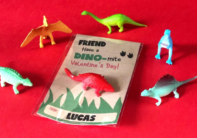 May include: A Valentine's Day card with a dinosaur theme. The card is brown with green grass at the bottom and says "Friend Have a Dino-mite Valentine's Day!" with dinosaur footprints. The card is signed "From Lucas". There are six colorful plastic dinosaur toys around the card.